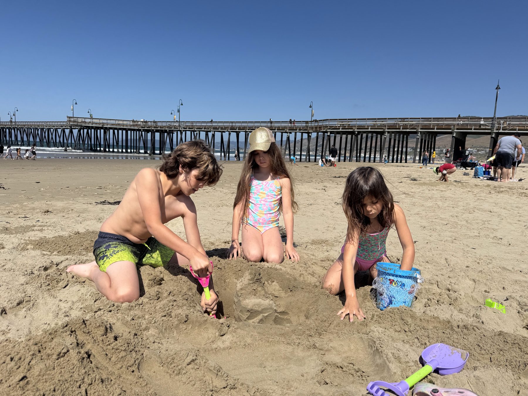 Three kids building sand castles with the Pismo pier behind them