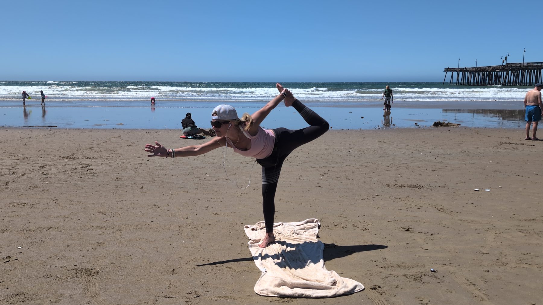 Dana doing Natarajasana dancer pose on the beach with the pier behind her